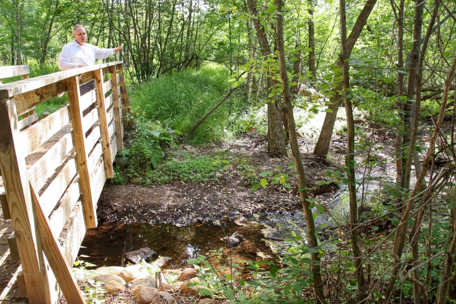 A man stands on a wooden bridge over a small creek gesturing to the surroundings.