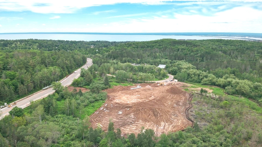 An aerial view of a construction area in the woods next to a highway with a large body of water on the horizon.