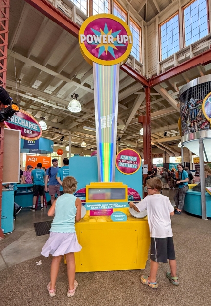 Two children spinning wheels on an interactive exhibit to power up lights in a column and illuminate the top sign that says "Power Up!"