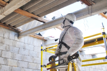 Worker in protective clothing and mask looks up at partially dismantled ceiling.
