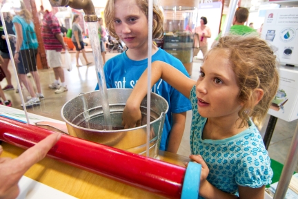 Girl puts hand in bucket of water at a water exhibit as another girl looks on.