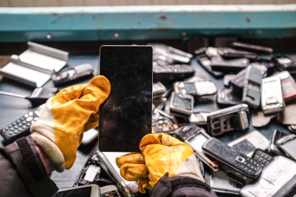 Hands wearing gloves holding discarded cell phone over a pile of old electronic devices.