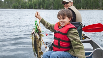 A smiling boy sitting in a boat on a lake holding up a string of walleye