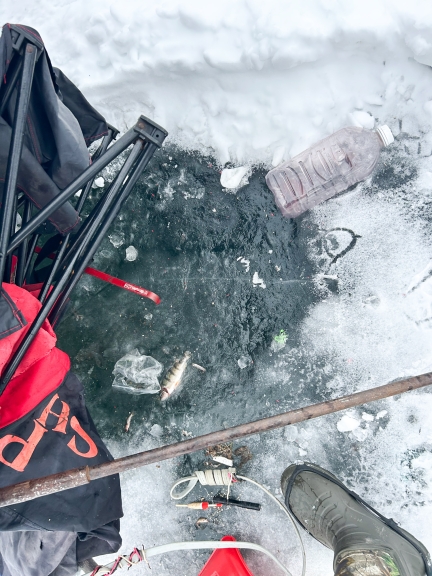 An  empty plastic bottle, a collapsed camp chair, and other garbage on the ice around an ice fishing hole.
