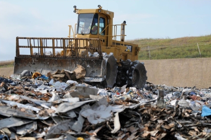 A yellow landfill compactor drives over trash in a landfill.