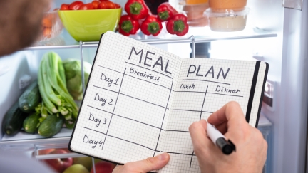 Closeup of hands holding a marker and a notebook with meal plan grid in front of an open refrigerator.