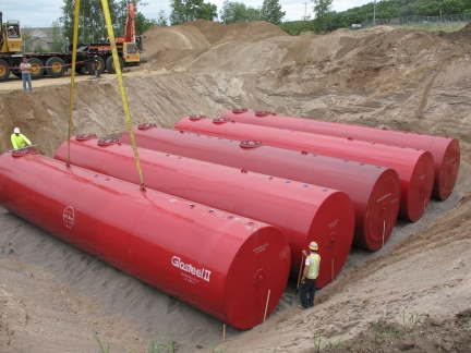 Workers installing five large red metal storage tanks in a freshly dug pit in the ground.