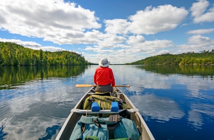 Woman wearing red shirt and hat sits in front of canoe on beautiful lake in wilderness.