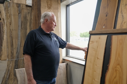 A man in a black t-shirt stands by a window looking at a piece of wood.