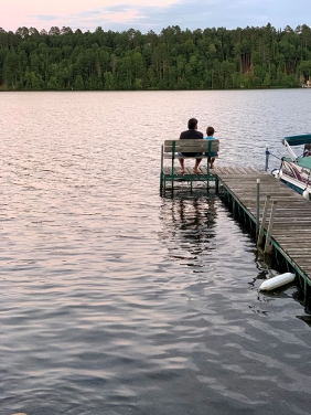 An adult and child sit on a bench the end of a dock, watching the sunset over the lake.