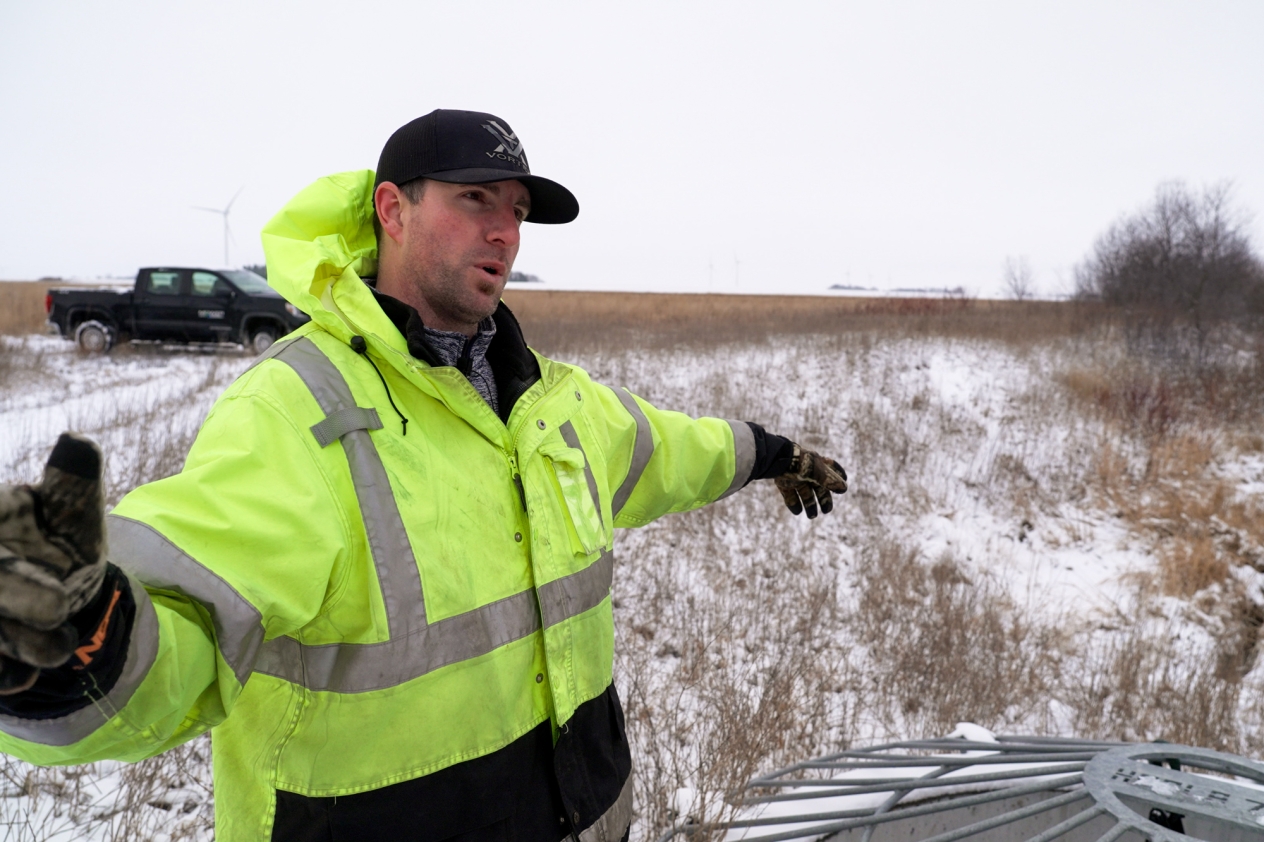 A man wearing a reflective jacket and black cap standing in a field during winter, talking and gesturing.