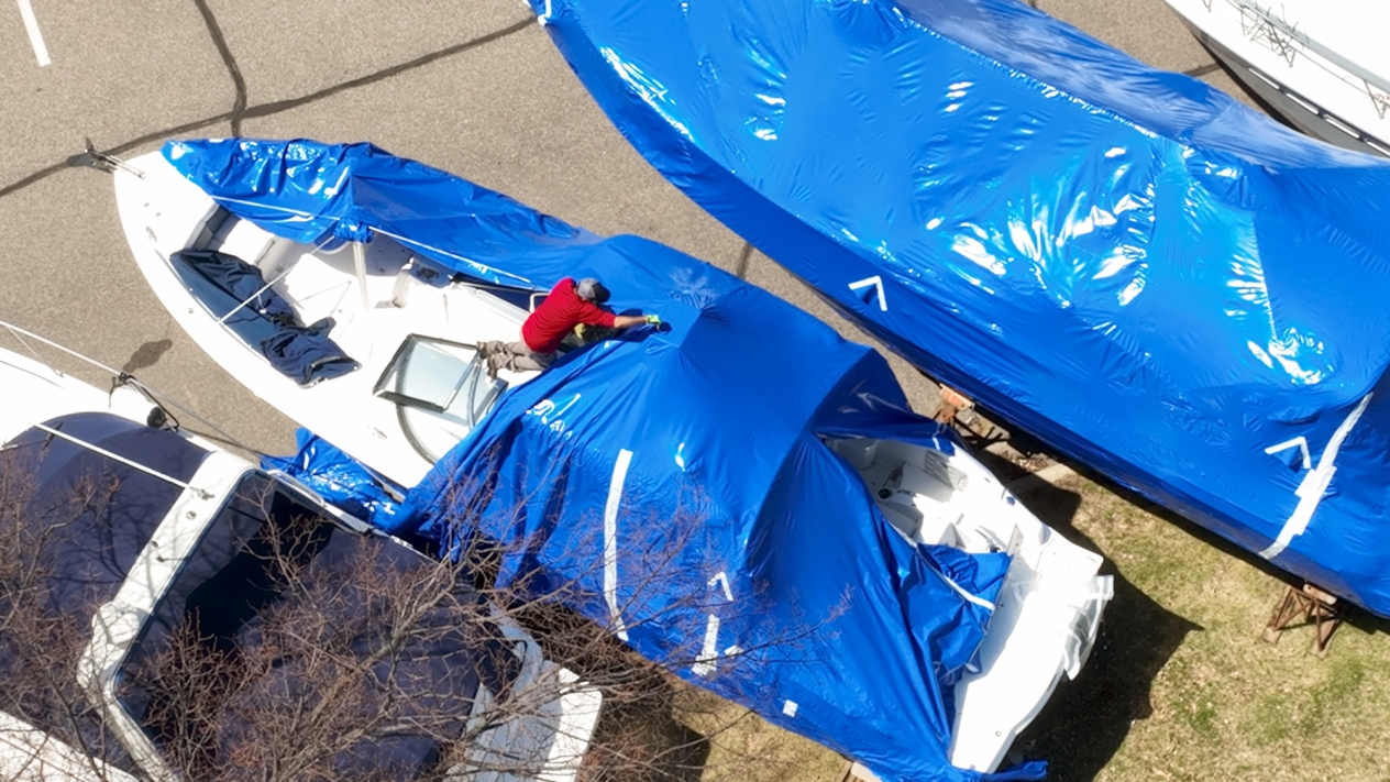 Aerial view of a man on a white boat in a parking lot removing blue boat wrap. 