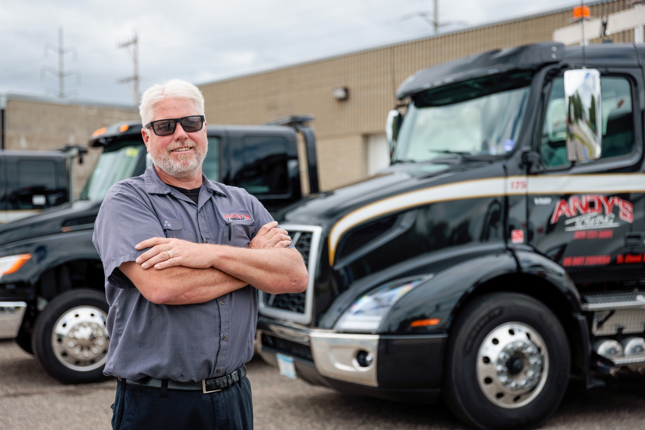 A smiling man with white hair and sunglasses stands in front of a black heavy-duty truck that says Andy's Towing on the door.