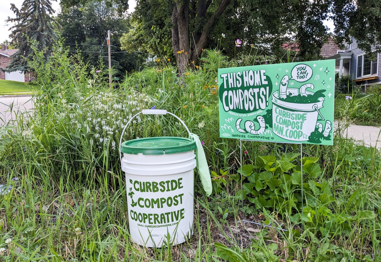 A bucket and sign for Curbside Compost Cooperative in a grassy area. The sign reads "This home composts! You can too."