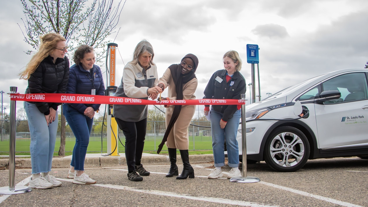 Officials cutting red ribbon in a parking lot to celebrate the opening of an electric vehicle charging station in St. Louis Park, Minnesota. From left: Ellie Rabine, sustainability specialist; Emily Ziring, sustainability manager; Sue Budd, ward 3 city council member; Mayor Nadia Mohamed; and Annie Pottorff, sustainability specialist.