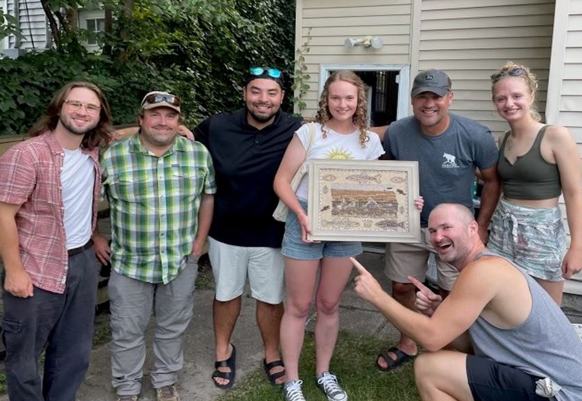 A group of young people smiling at the camera and displaying a framed piece of seed art.