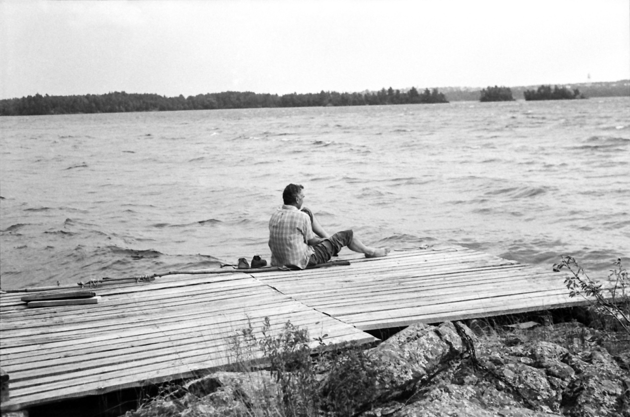 Black and white photo of a man sitting next to a lake on a wooden platform.