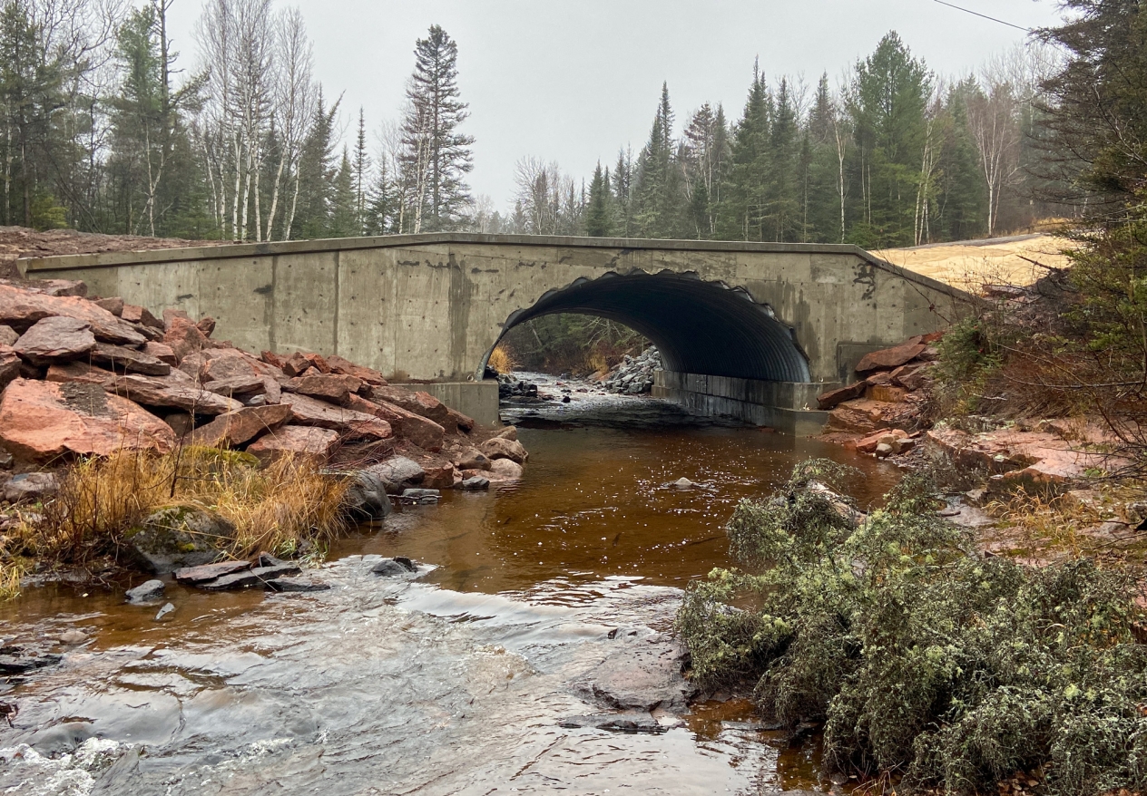 A cement bridge over a brown stream with a large culvert through it.