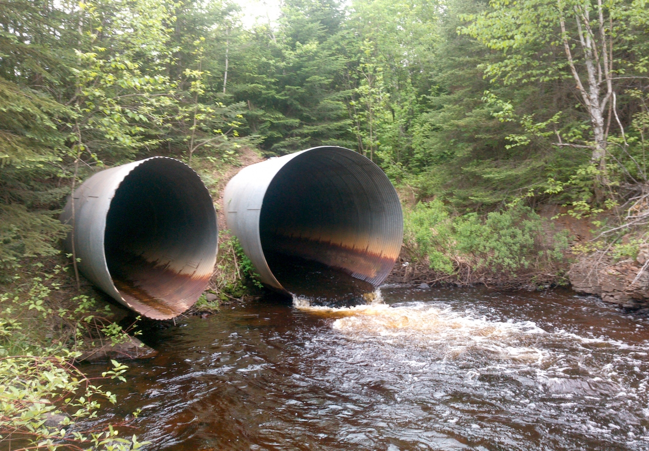 Two large corrugated metal pipes sticking out from a hill with water running out of the pipe on the right.