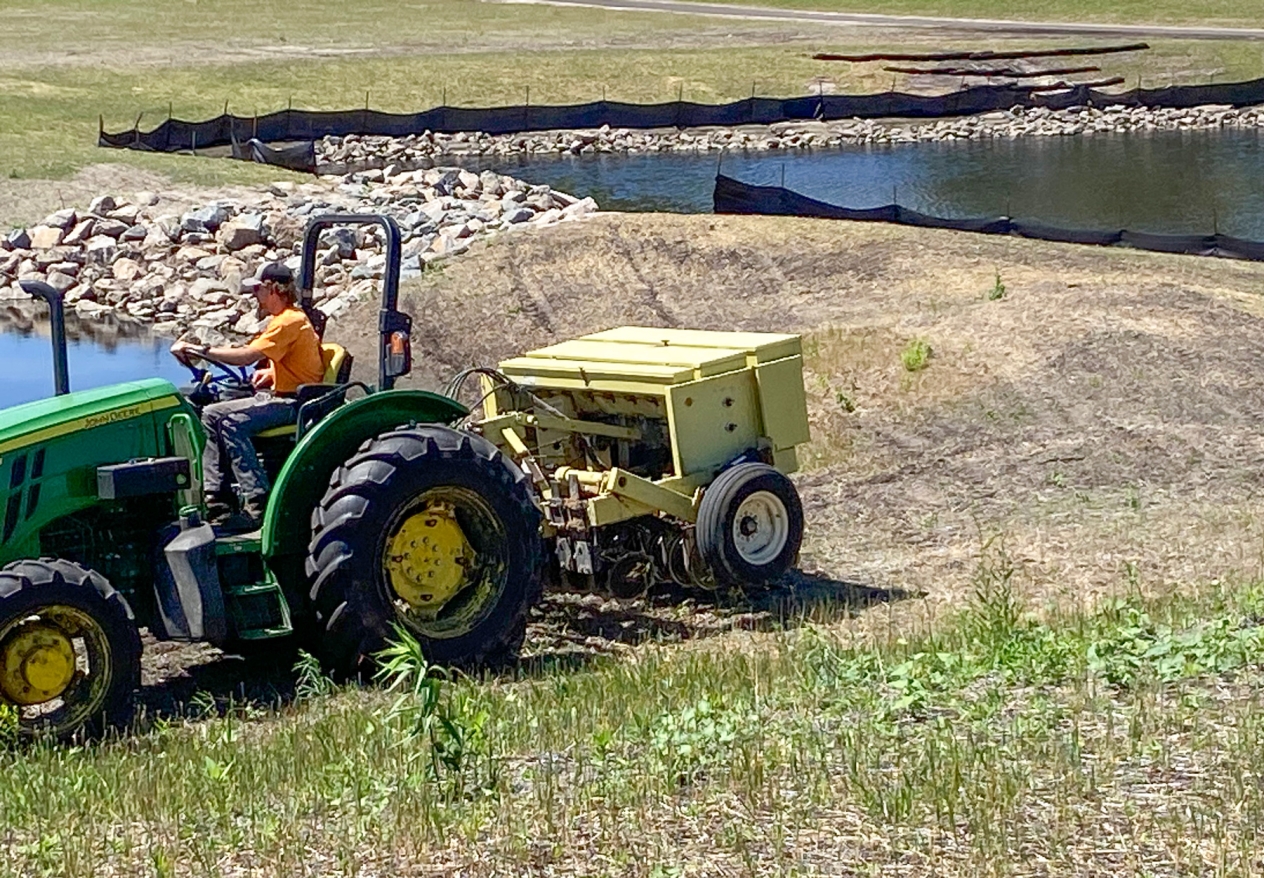 A worker driving a small John Deere tractor pulling a no-till seed planting drill next to stormwater ponds.