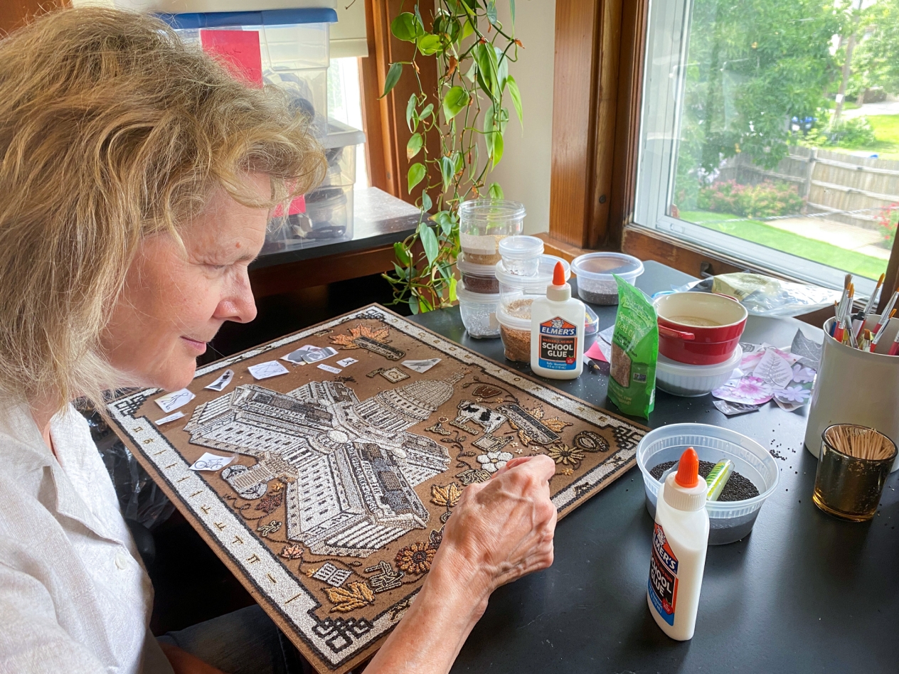 A woman sitting at a table with bottles of glue and containers of various seeds working on a picture made of seeds.