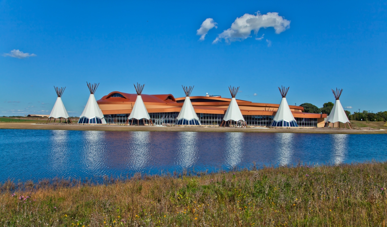 A low glass and earth-colored building behind a row of seven white tipis. A lake is in the foreground.